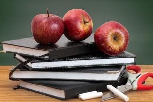 Four black books stacked with 3 apples on top of them placed on a table in front of a chalkboard. 